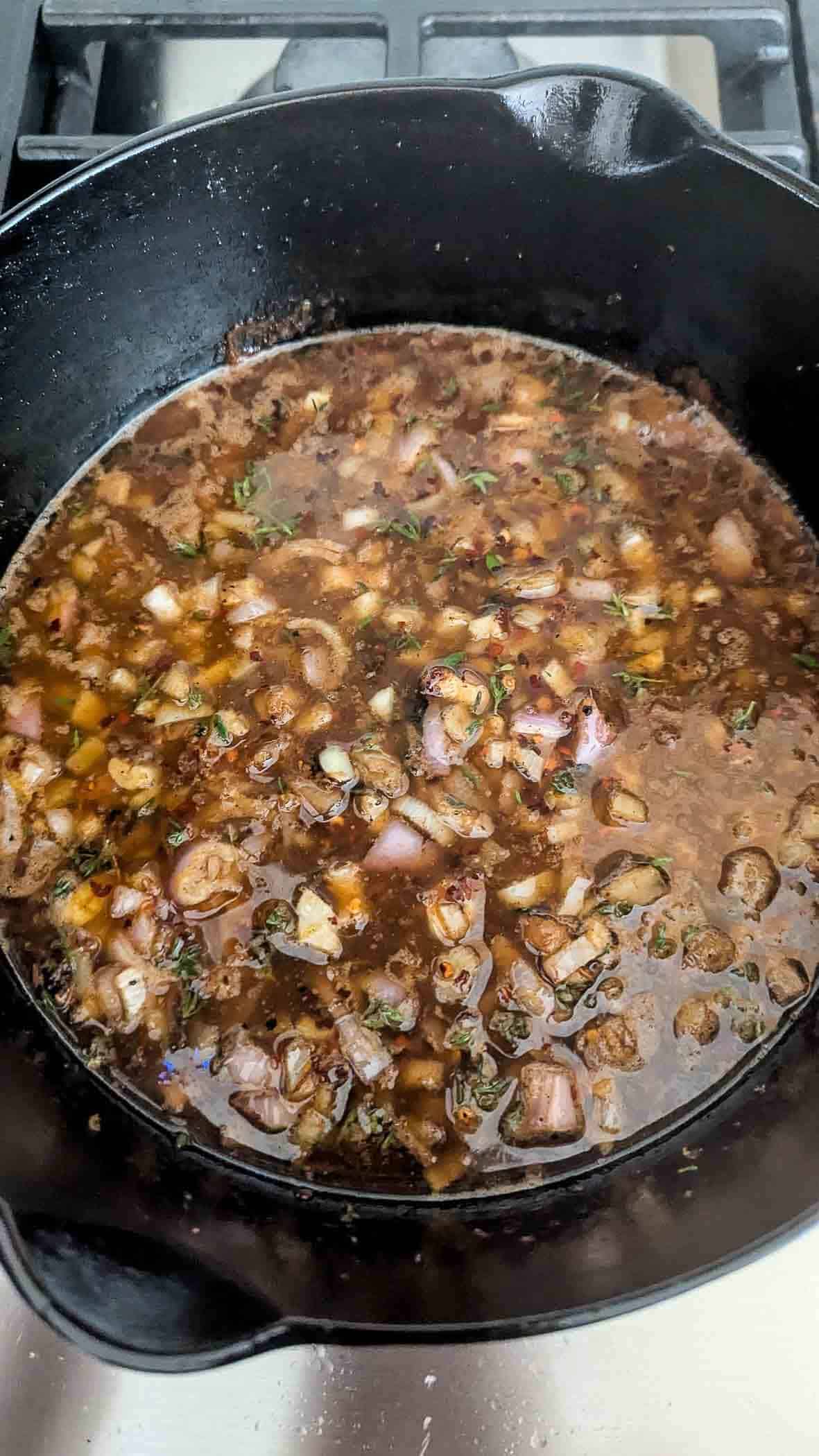 A cast iron skillet on a stovetop filled with a simmering mixture of chopped onions, herbs, and a dark, flavorful liquid.