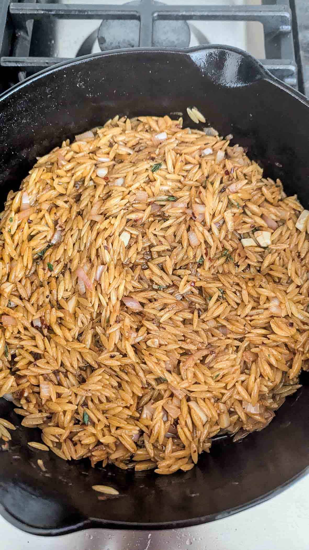 A cast iron skillet on a stove contains browned orzo pasta mixed with chopped onions and herbs, ready to be cooked further.