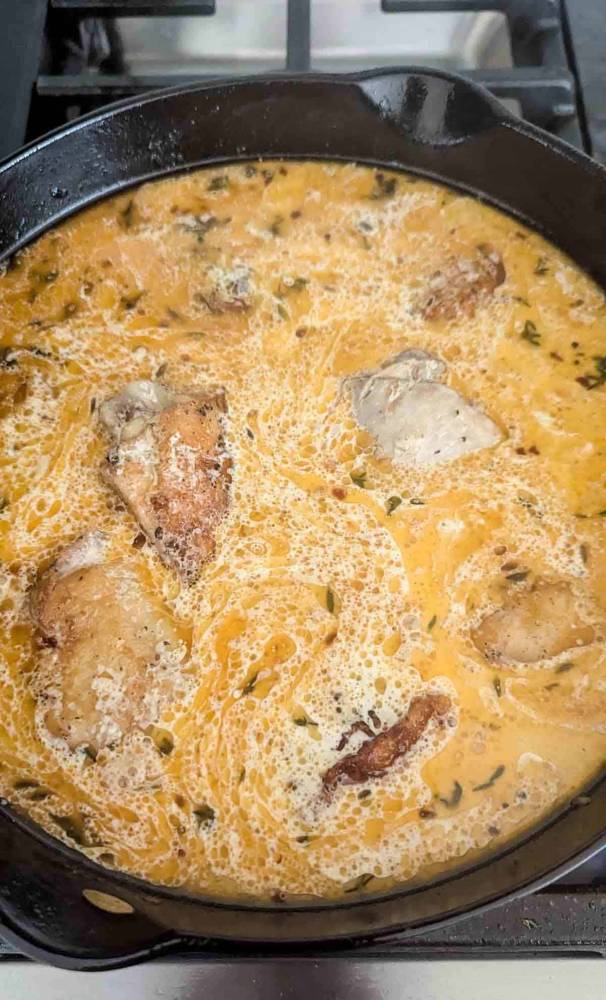 Chicken pieces simmering in a creamy sauce with herbs, cooking in a black cast iron skillet on a stovetop.