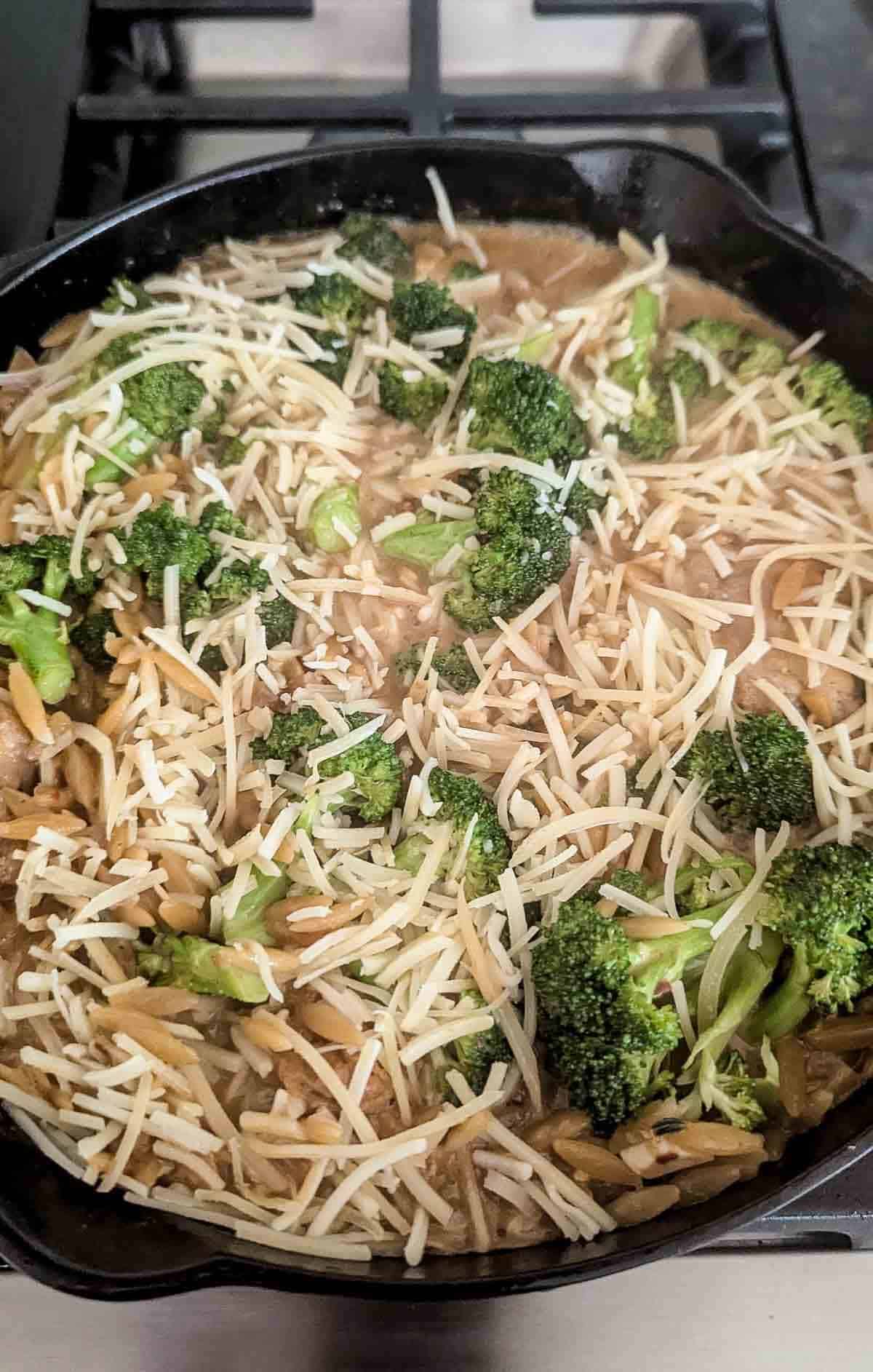 A skillet filled with a creamy chicken and orzo dish, topped with shredded cheese and broccoli florets, sits on a stove.