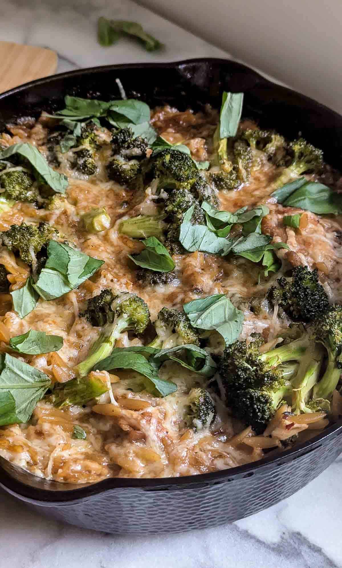 A lemon chicken orzo skillet, topped with roasted broccoli, melted cheese, and fresh basil leaves, sits on a marble countertop.