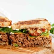 A close-up of a sliced meatloaf sandwich on toasted bread, with layers of lettuce, tomato, cheddar cheese, and leafy greens, displayed on a wooden cutting board.