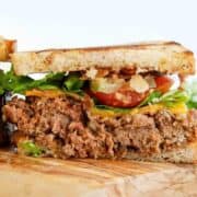 Close-up of a sandwich with toasted bread, a thick slice of meatloaf, lettuce, tomato slices, cheddar cheese, and crispy bacon, resting on a wooden surface.