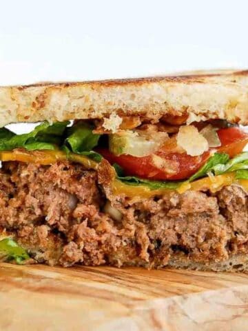 Close-up of a sandwich with toasted bread, a thick slice of meatloaf, lettuce, tomato slices, cheddar cheese, and crispy bacon, resting on a wooden surface.