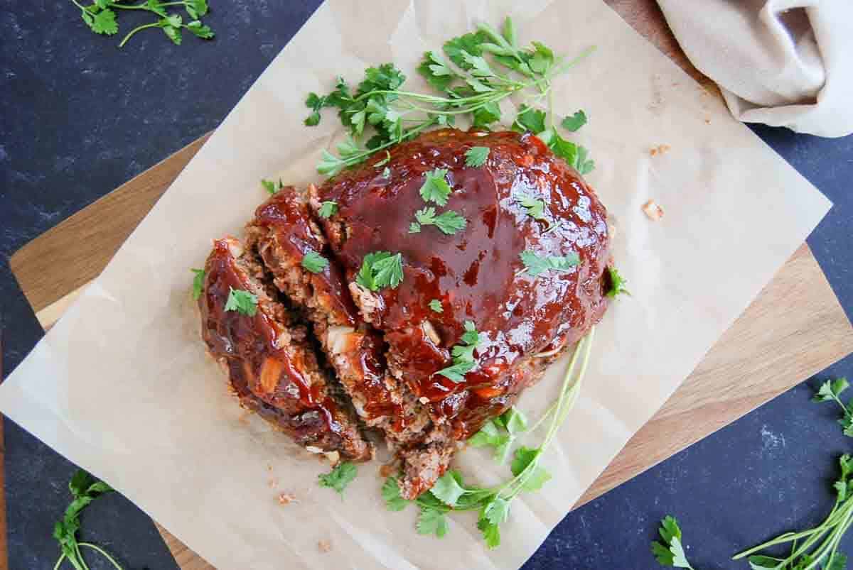 A glazed meatloaf topped with fresh parsley is sliced and served on parchment paper atop a wooden board, with extra parsley scattered around.
