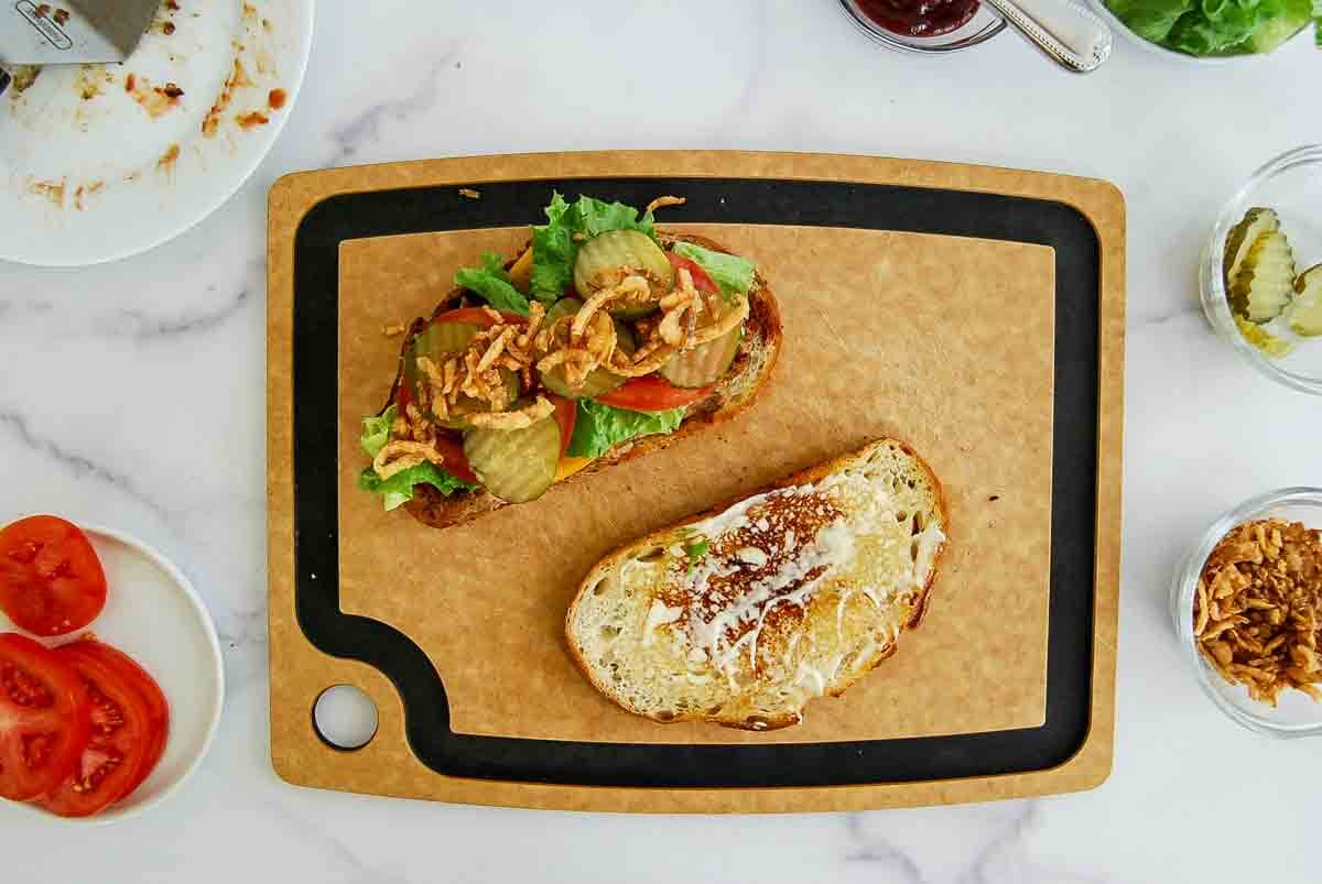 An open face meatloaf sandwich on a cutting board, topped with lettuce, pickle slices, tomato, and crispy onions. The other slice of bread is spread with mayonnaise. Nearby are bowls with more pickles, crispy onions, and sliced tomatoes.