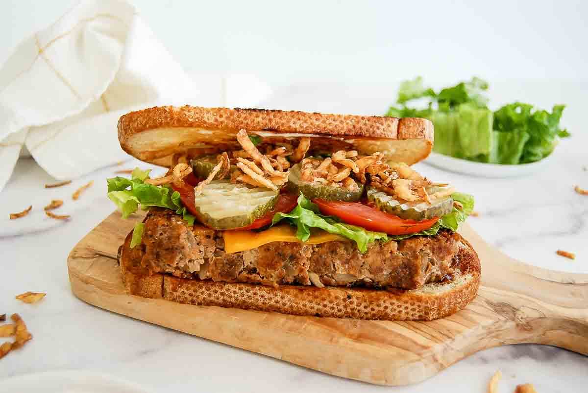 A hearty sandwich with meatloaf, cheddar cheese, lettuce, tomato, pickles, jalapeños, crispy fried onions, and toasted bread sits on a wooden cutting board. A napkin and lettuce are in the background.