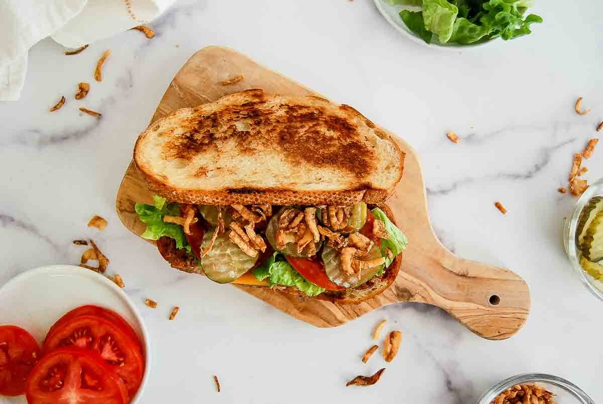 A toasted meatloaf sandwich with lettuce, tomato, pickles, and crispy onions sits on a wooden cutting board, surrounded by bowls of sliced tomatoes, pickles, and lettuce on a white marble surface.