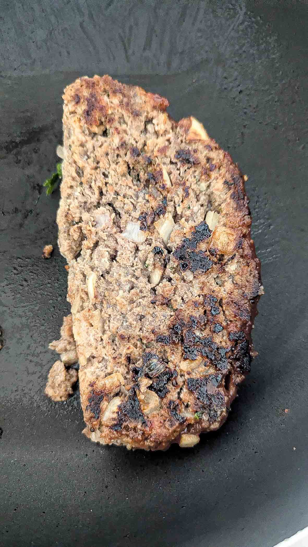 A cooked slice of meatloaf with visible chunks of onions sits on a dark, flat surface, showing a browned and slightly charred exterior.