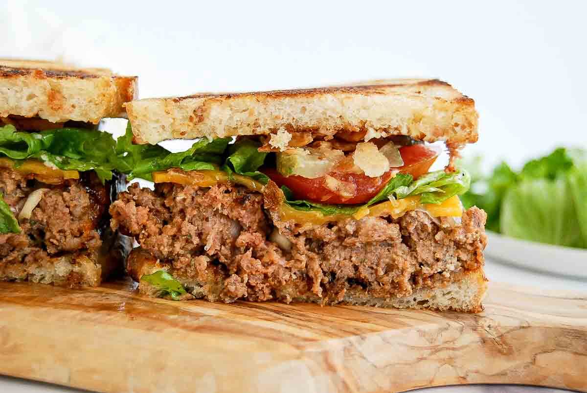 A close-up of a sliced meatloaf sandwich on a wooden board, with cheddar cheese, lettuce, tomato, and crispy bacon, between toasted bread slices.