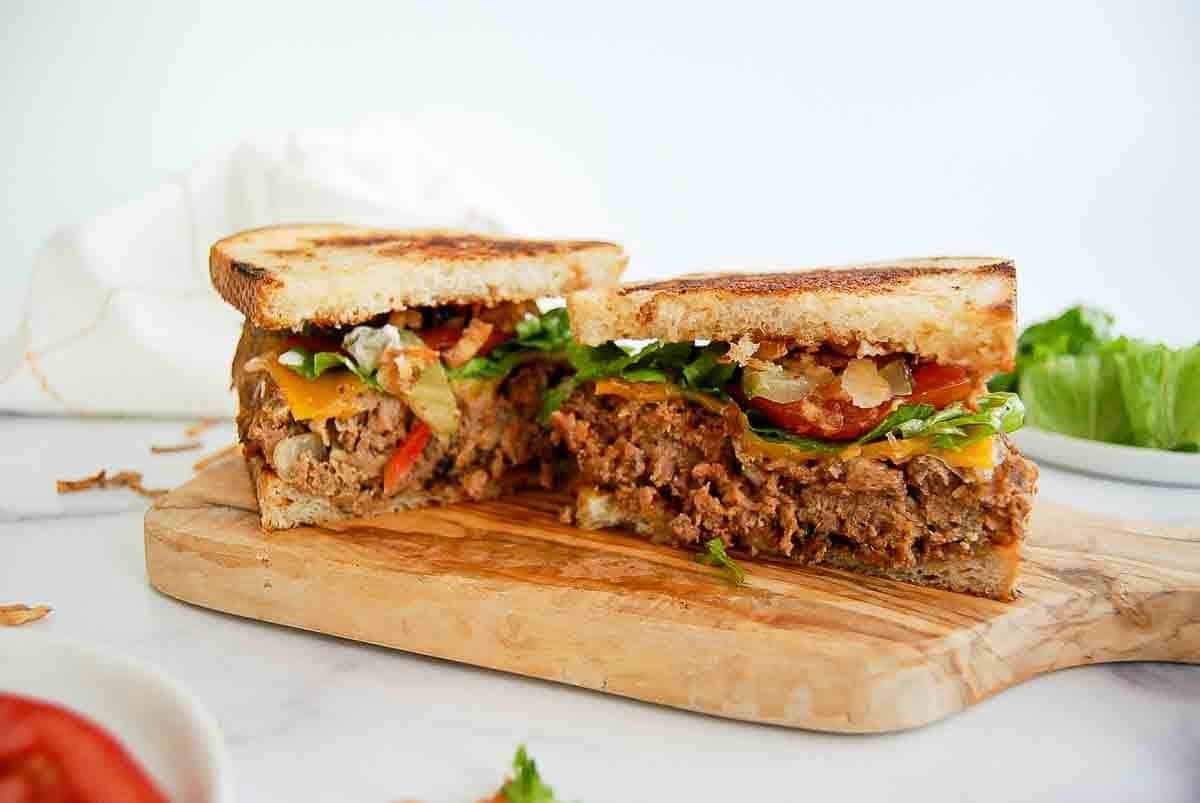 A sliced meatloaf sandwich with lettuce, tomato, and grilled onions on toasted bread sits on a wooden cutting board, with fresh lettuce and tomato visible in the background.