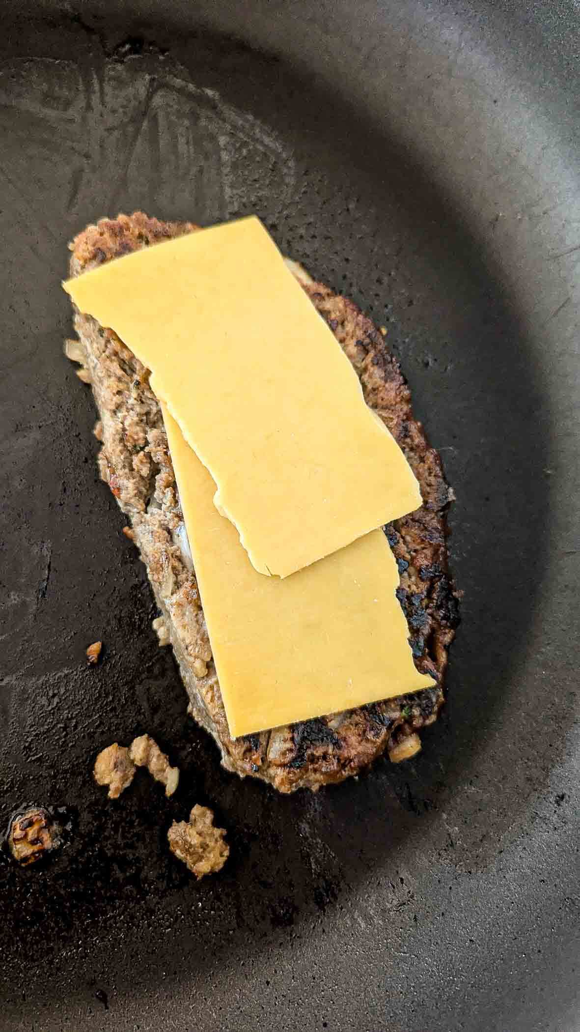 A meatloaf slice in a skillet with two uneven slices of yellow cheese placed on top, starting to melt.
