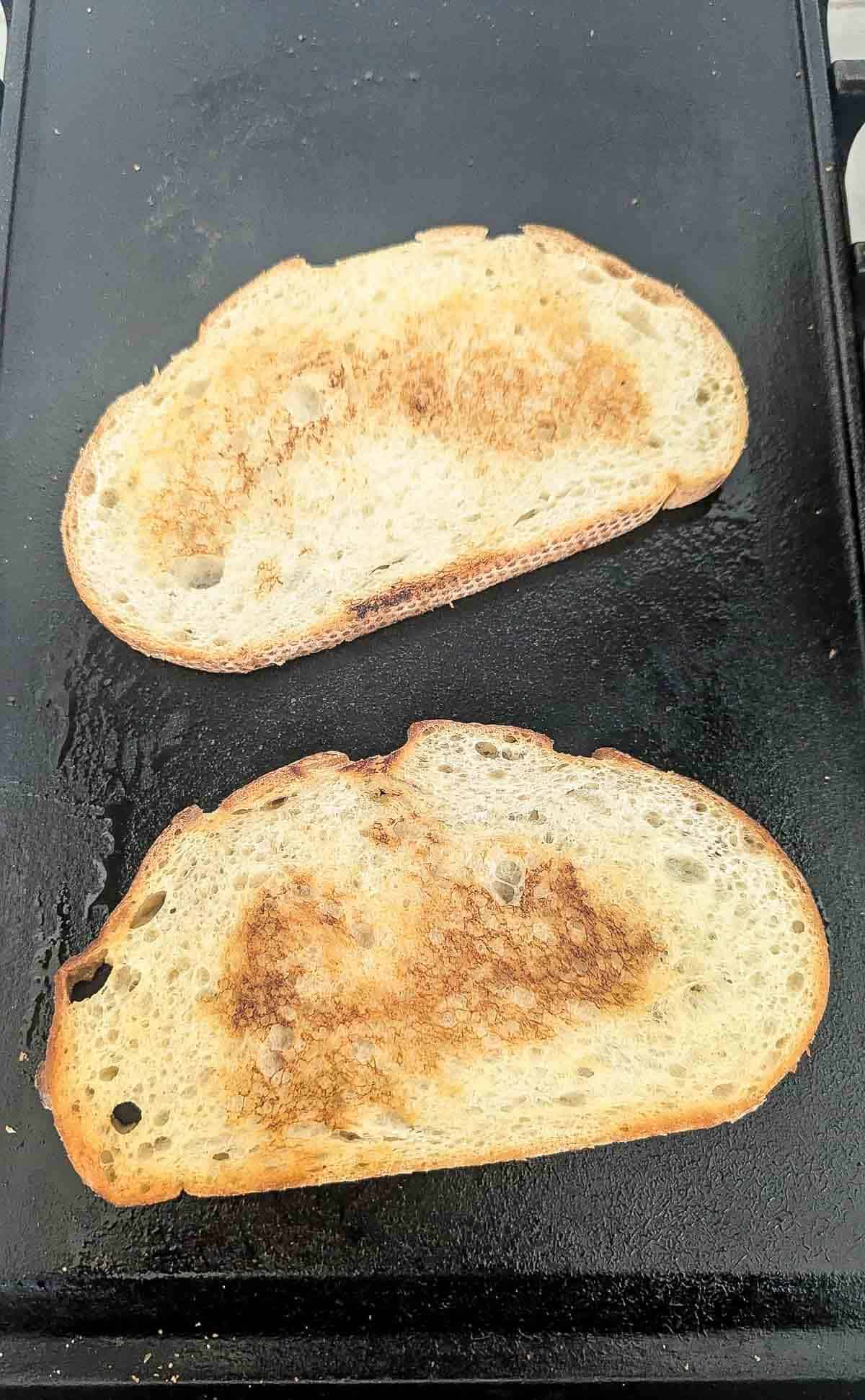 Two slices of bread toasting on a black griddle, both showing golden brown spots and uneven toasting.