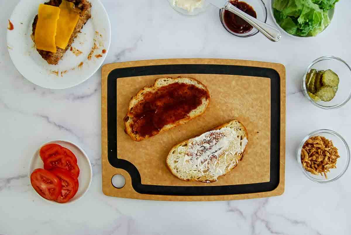 Two slices of bread on a cutting board, one with mayonnaise and the other with barbecue sauce, surrounded by plates of sliced tomato, pickles, cheese-topped meatloaf, crispy onions, lettuce, and sauce bowls.