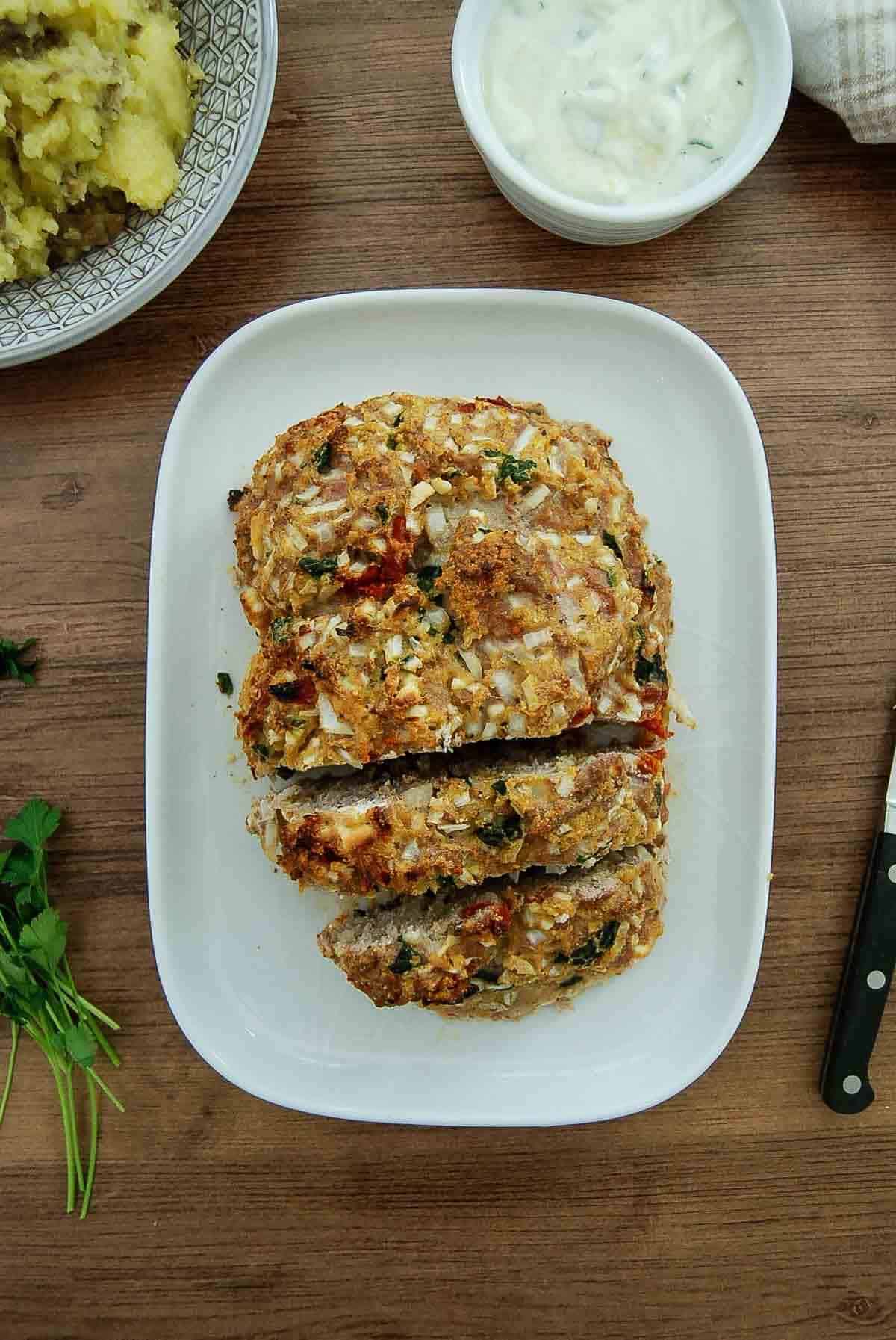 A loaf of baked greek turkey meatloaf with chopped herbs, topped with onions, is sliced on a white platter.