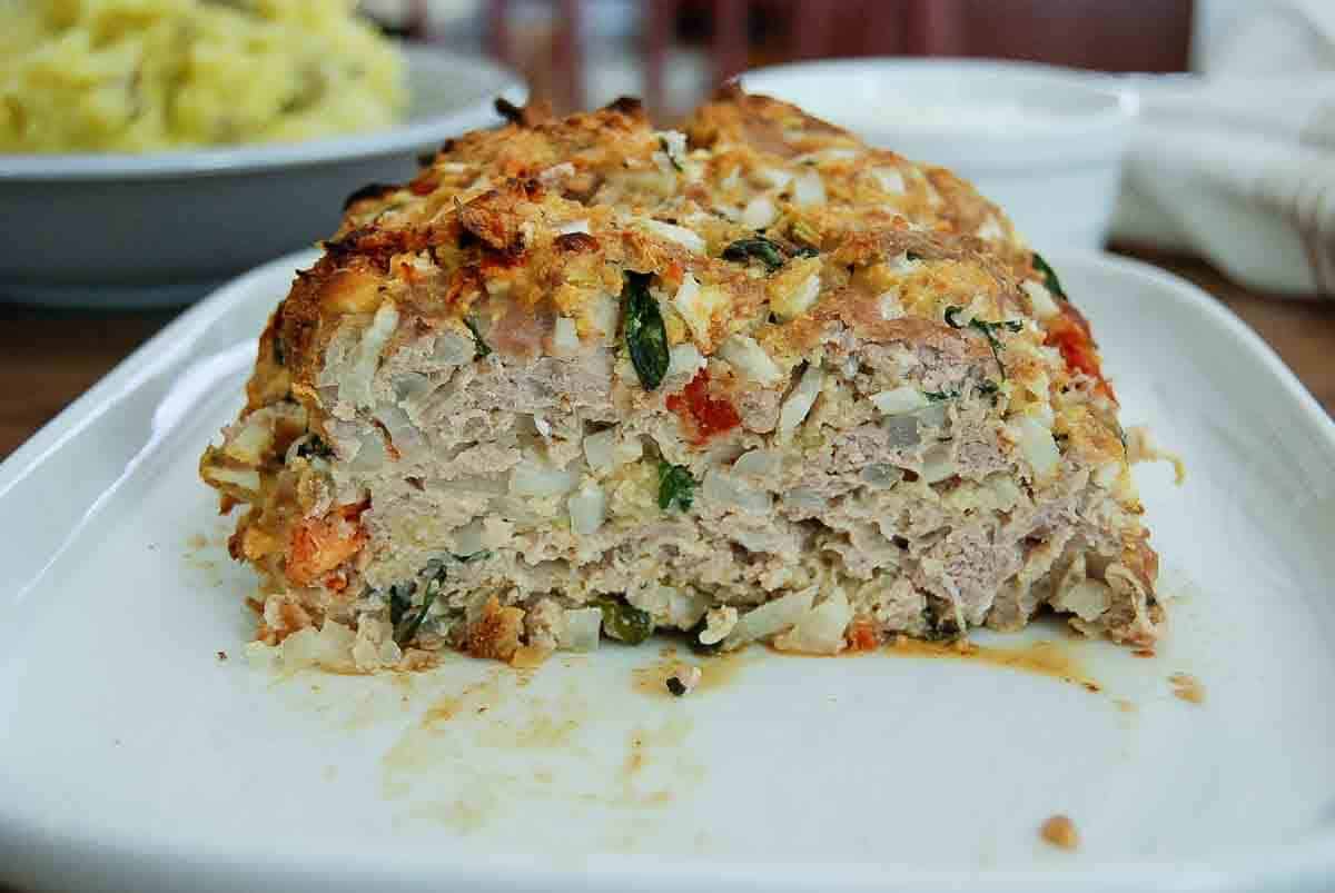 A close-up of a sliced turkey meatloaf on a white platter, showing its moist interior mixed with vegetables and herbs.