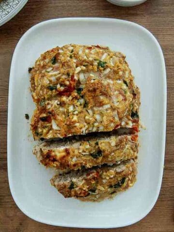 A loaf of homemade greek turkey meatloaf topped with chopped onions and herbs, partially sliced and served on a white rectangular plate, with a parsley garnish and knife placed nearby on a wooden table.