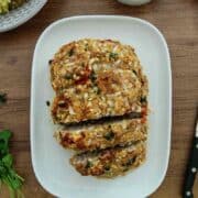 A loaf of cooked turkey meatloaf with herbs, sliced on a white plate, sits on a wooden table next.