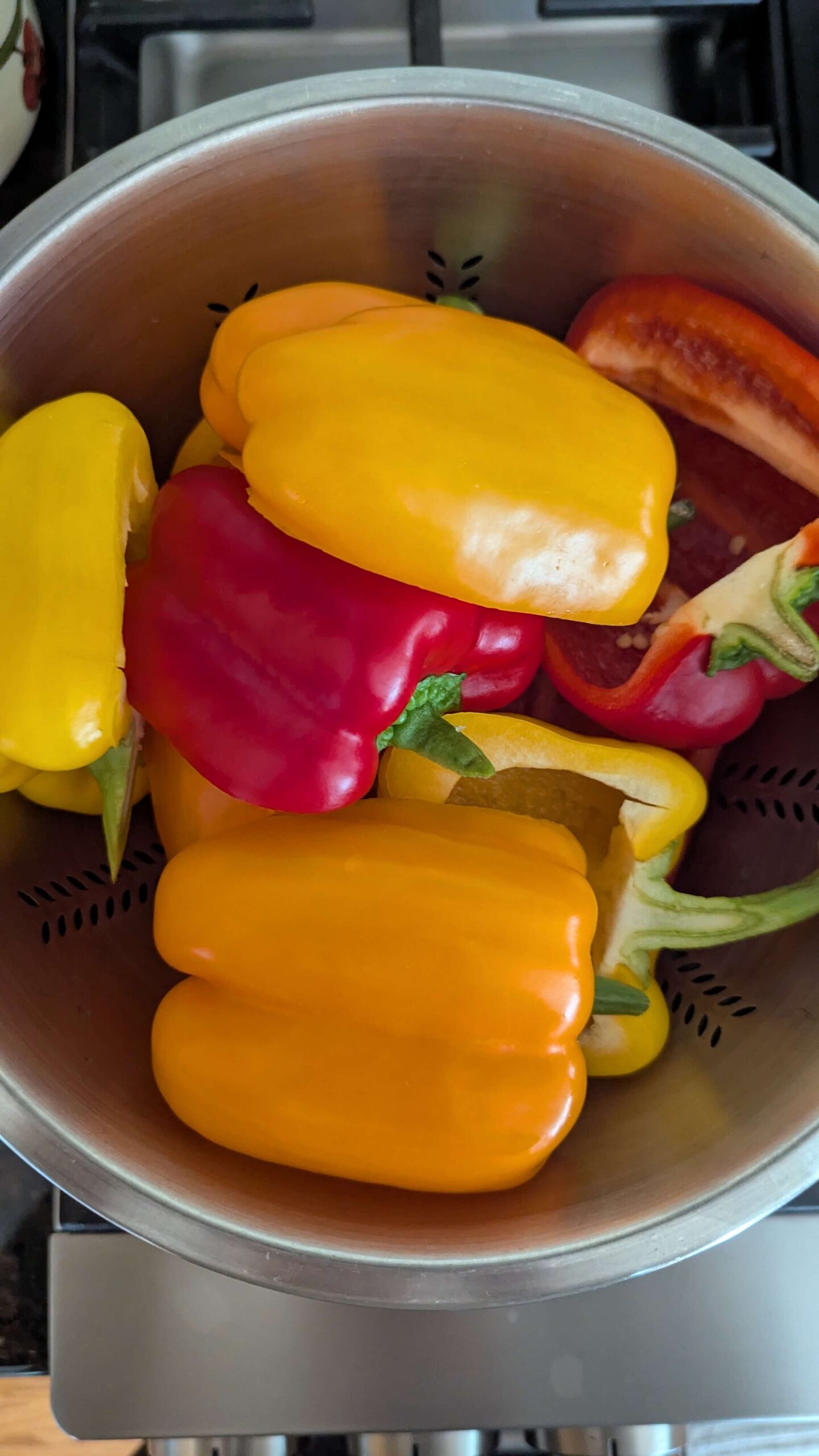 A metal colander filled with whole yellow, orange, and red bell peppers, some with stems attached, sits on a stovetop.