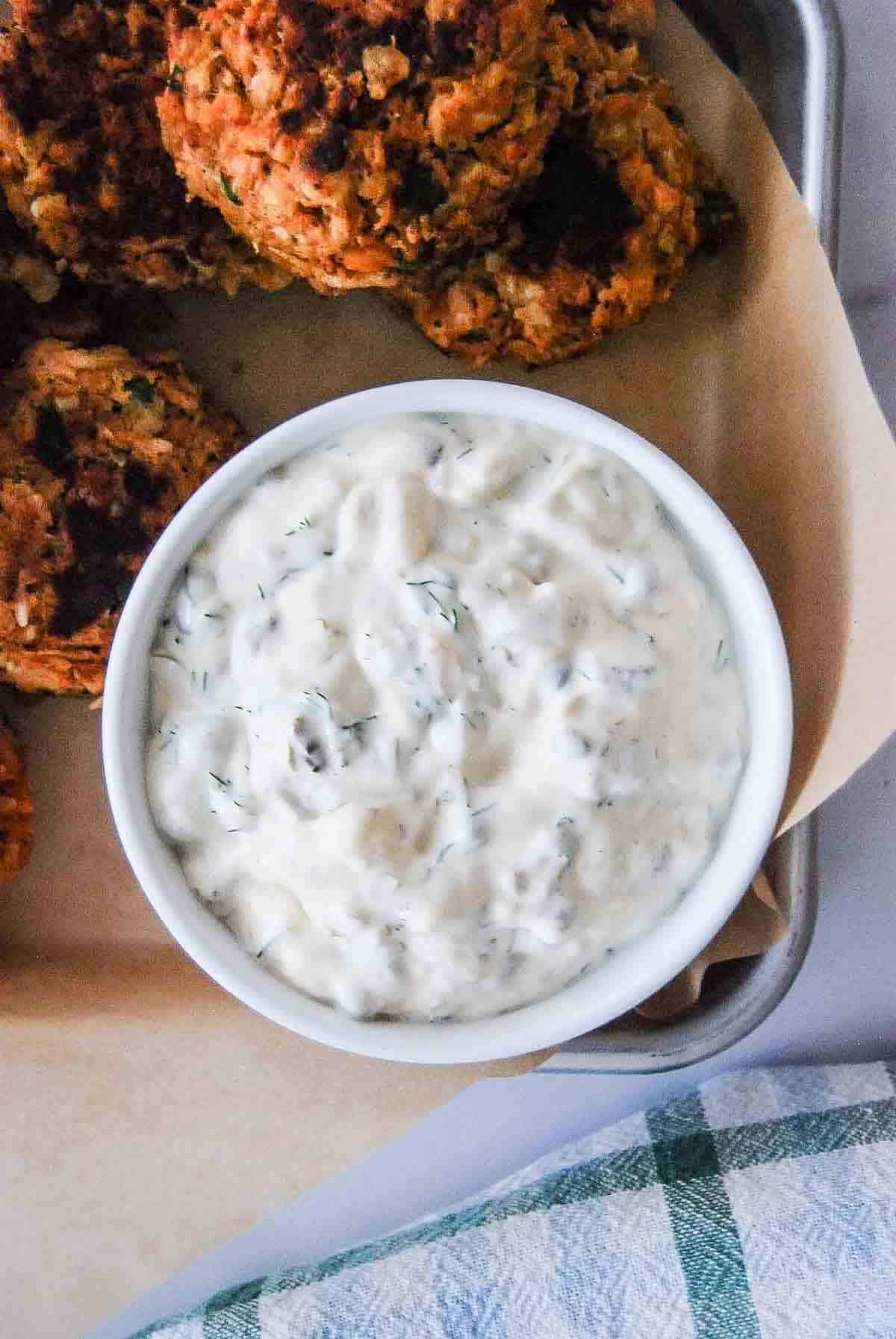A white bowl filled with creamy homemade tartar sauce with herbs sits on a tray next to several golden-brown salmon cakes.