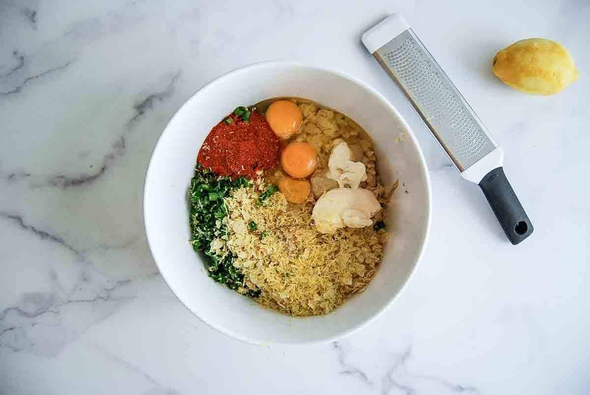 A white bowl on a marble surface filled with eggs, chopped herbs, spices, grated lemon zest, and breadcrumbs. Nearby are a whole lemon and a grater.