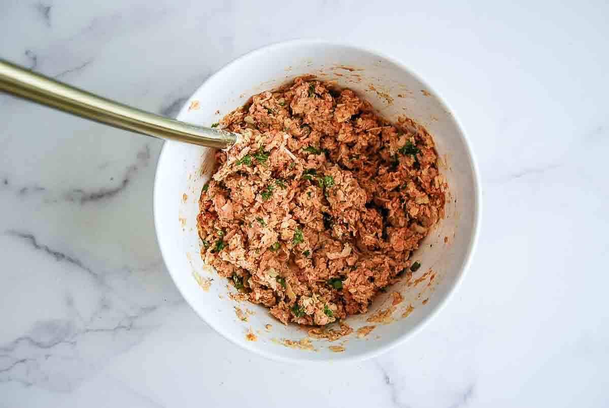 A white bowl with a metal spoon holds a mixture of ground meat, herbs, and spices on a white marble surface.
