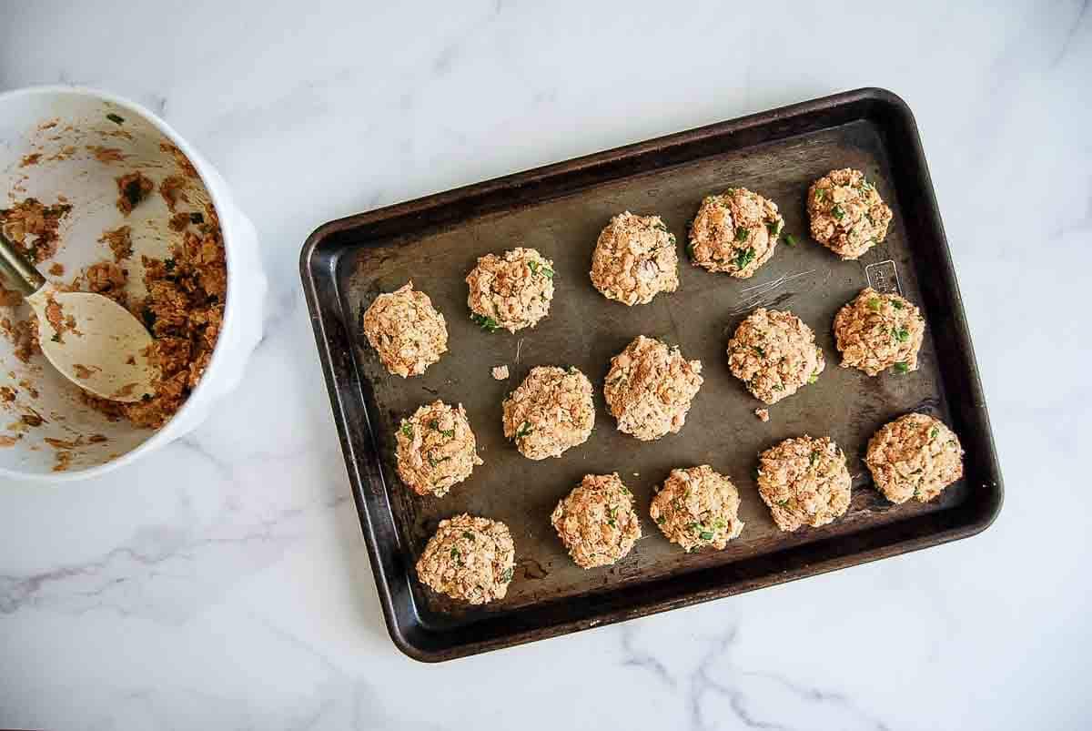 A baking tray with twelve uncooked, round balls of mixed ingredients sits on a marble surface. Next to it is a mixing bowl with leftover mixture and a spatula inside.