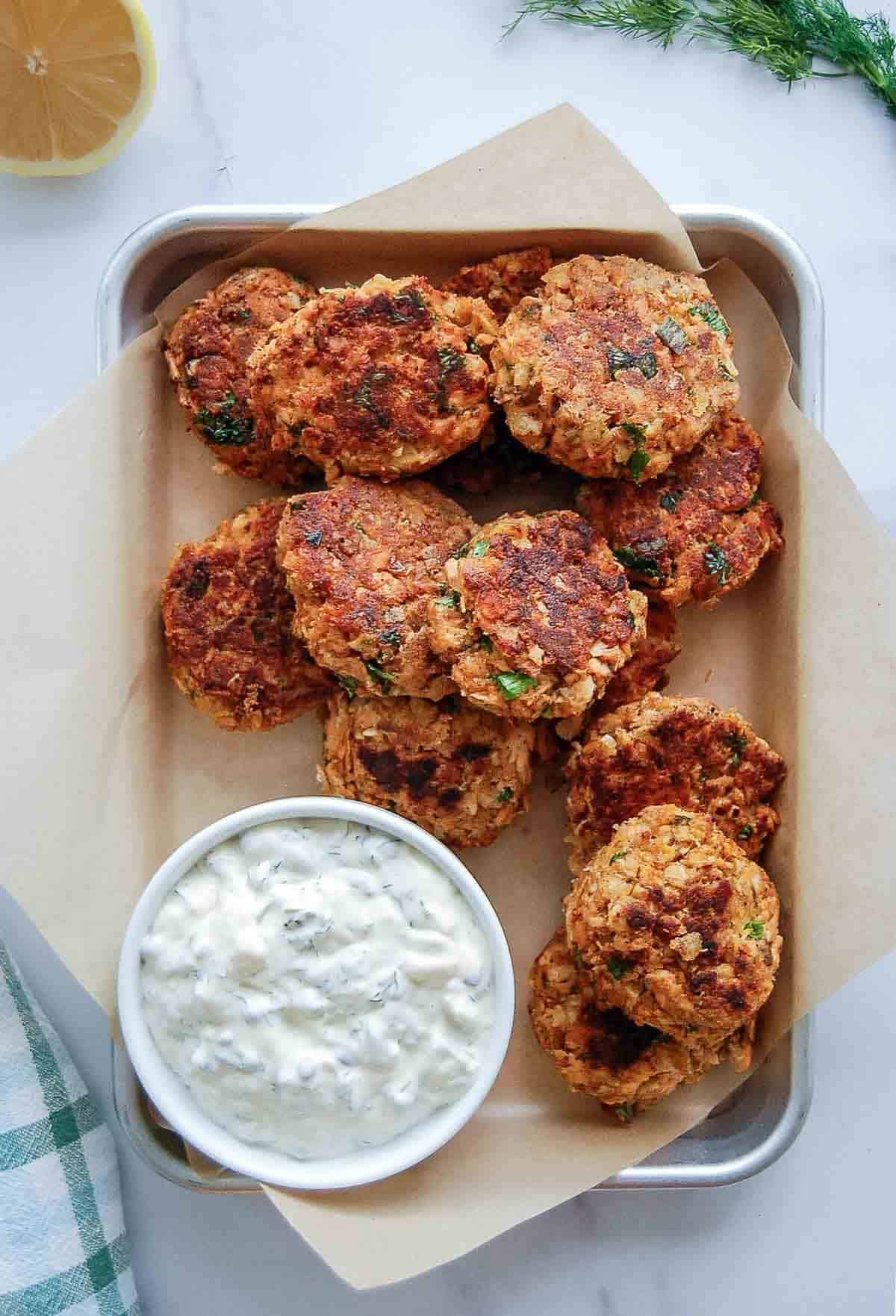 A metal tray lined with parchment holds several golden-brown salmon patties and a bowl of creamy tartar sauce with herbs. A lemon wedge and fresh dill are nearby on a white surface.