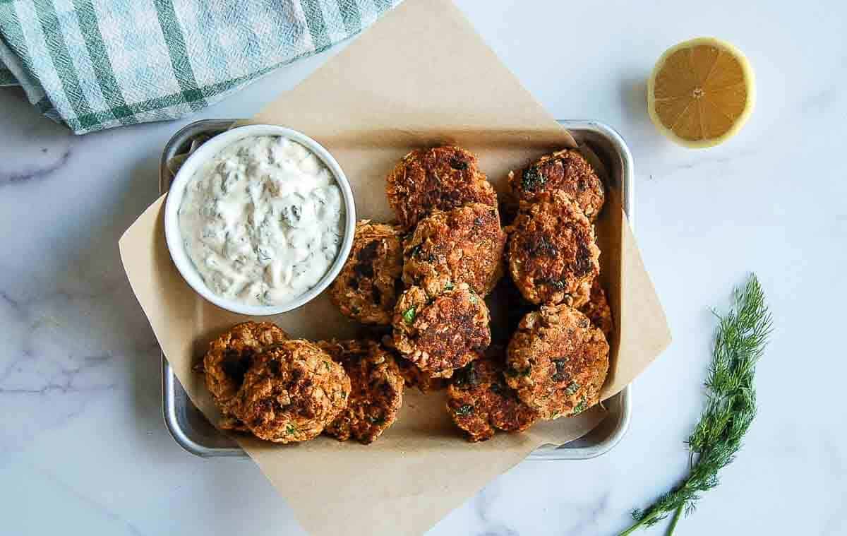 A tray lined with parchment paper holds several golden brown salmon fritters beside a bowl of creamy tartar sauce. A halved lemon, fresh dill, and a green-checked towel are nearby on a marble surface.