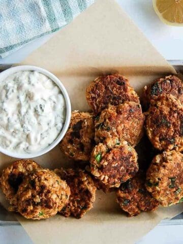 A tray lined with parchment paper holds several golden brown salmon fritters next to a bowl of creamy tartar sauce. A lemon wedge and kitchen towel are visible in the corner.