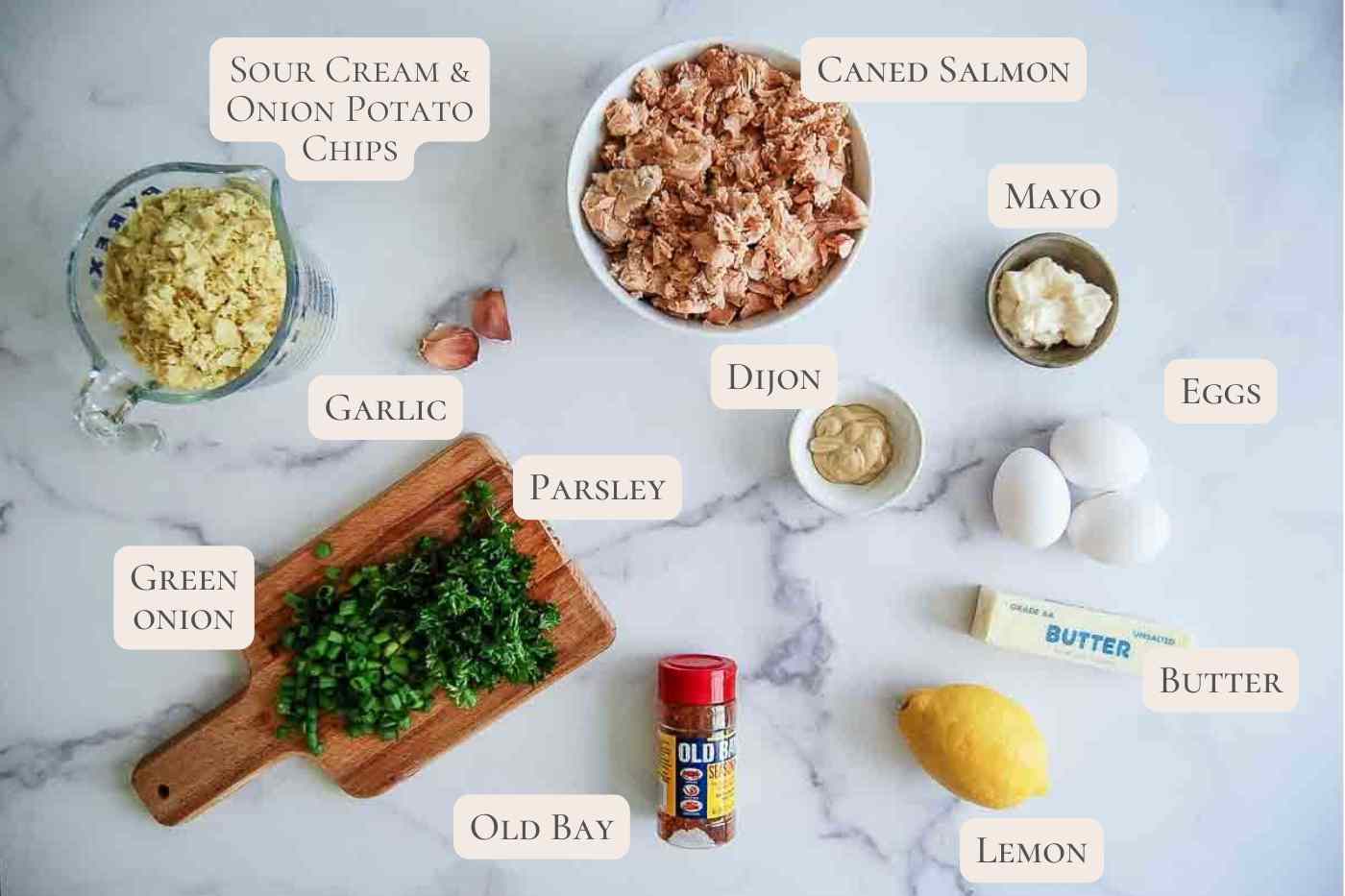 Ingredients for salmon cakes arranged on a white surface, including canned salmon, eggs, mayo, Dijon, butter, lemon, Old Bay, chopped parsley, green onion, garlic, and crushed sour cream & onion potato chips.