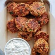 A tray lined with parchment paper holds several golden-brown salmon patties garnished with herbs, next to a bowl of creamy tartar sauce.