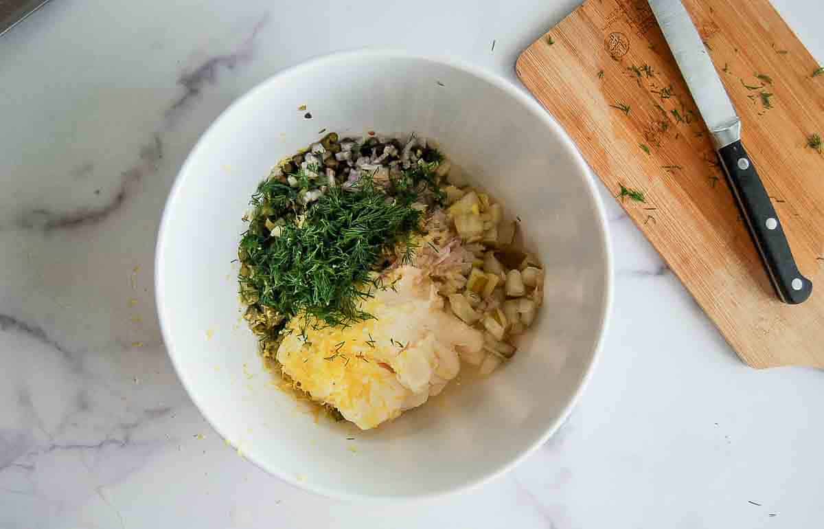 A white bowl on a marble counter contains chopped onions, and herbs, seasonings, next to a cutting board with a knife and scattered fresh dill.