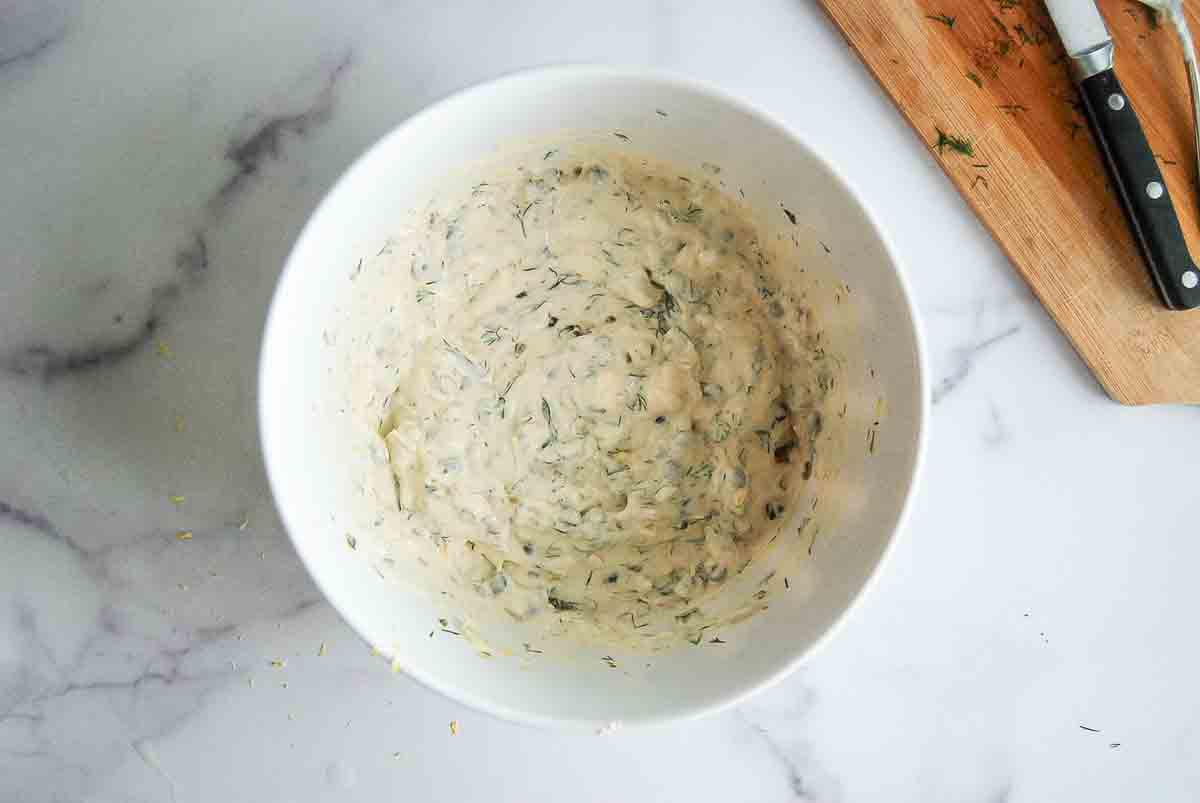 A white bowl filled with a creamy, herb-flecked sauce sits on a marble surface next to a wooden cutting board with a knife and scattered fresh herbs.