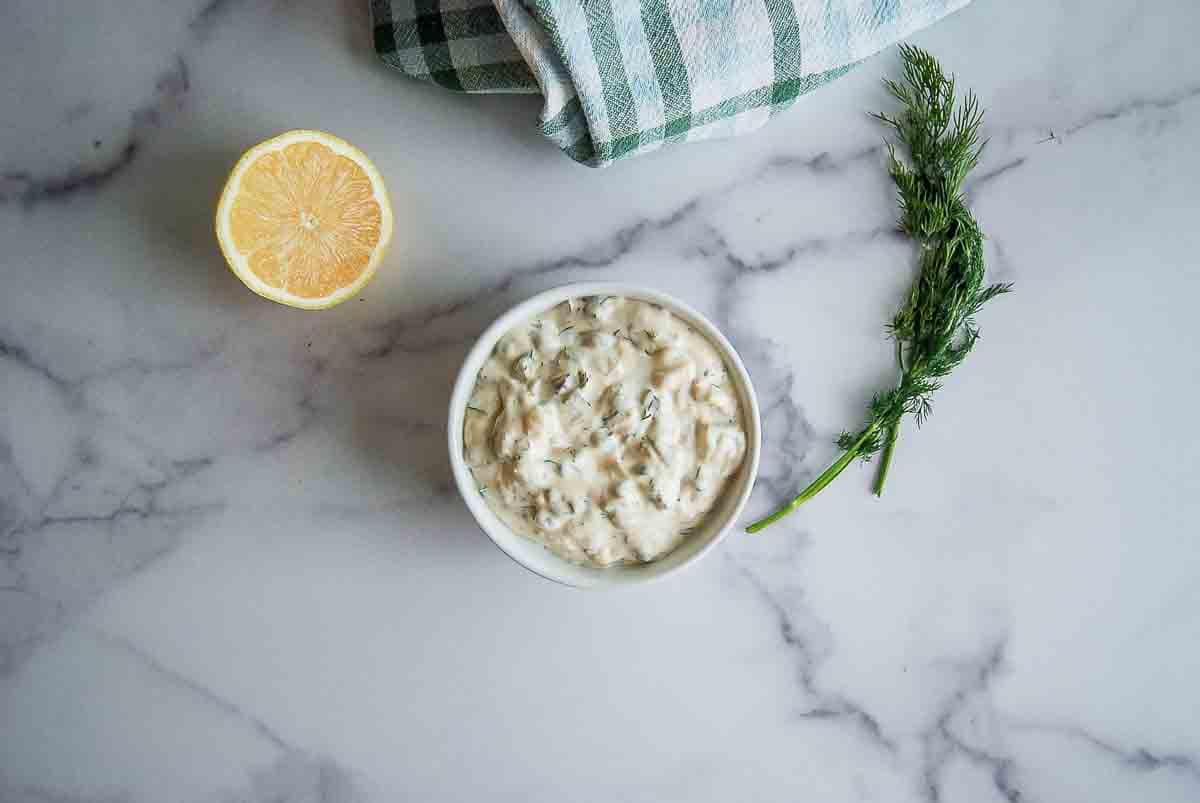 A bowl of creamy dip with herbs sits on a marble surface next to a half lemon, a sprig of dill, and a folded green-and-white checkered cloth.