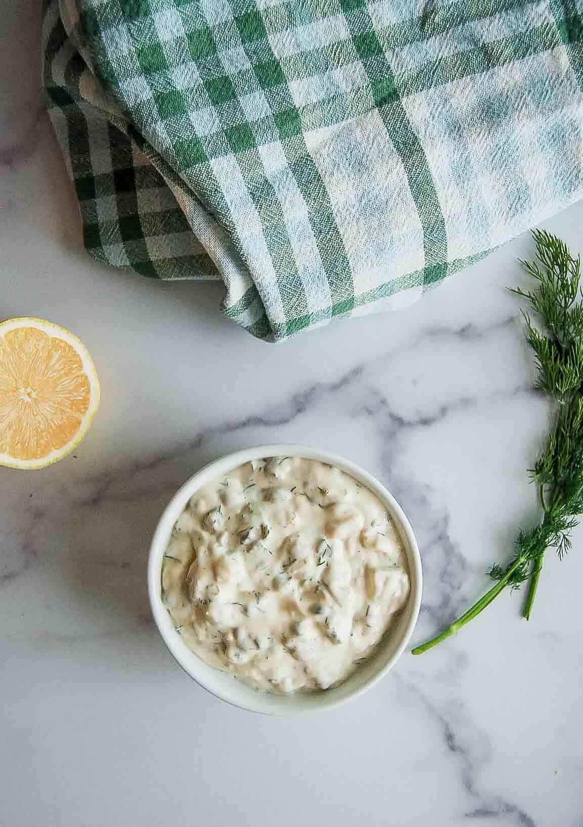 A bowl of creamy tartar with herbs sits on a marble surface next to a sprig of fresh dill, half a lemon, and a folded green-and-white checkered towel.