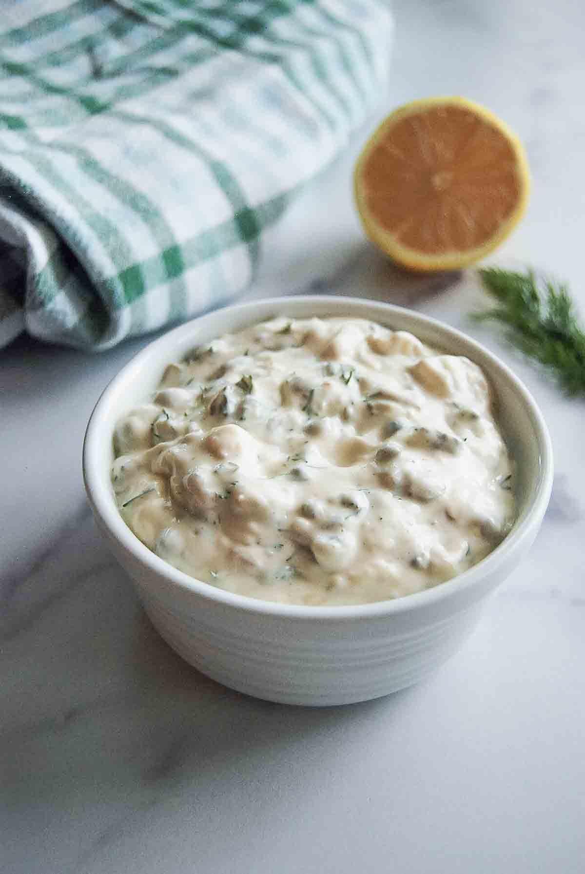 A white bowl filled with creamy tartar sauce with herbs sits on a white surface. A half lemon, a sprig of dill, and a green-and-white striped cloth are in the background.