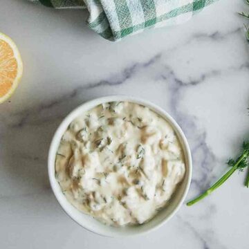 A small white bowl of creamy tartar sauce with chopped herbs sits on a marble surface, next to a lemon half, fresh dill, and a green and white checkered towel.
