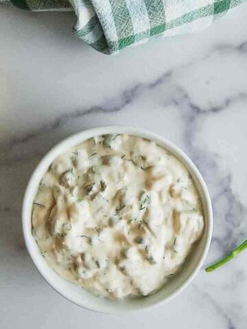 A small white bowl of creamy tartar sauce with chopped herbs sits on a marble surface, next to a lemon half, fresh dill, and a green and white checkered towel.