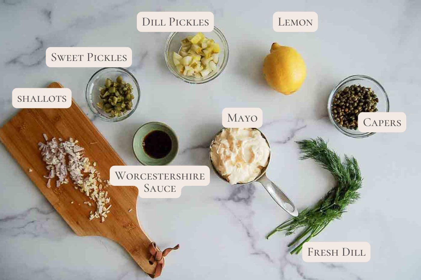 Ingredients for tartar sauce: chopped shallots on a cutting board, bowls of sweet and dill pickles, mayo, lemon, capers, Worcestershire sauce, and a sprig of fresh dill on a white surface.