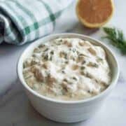 A white bowl filled with creamy tartar sauce sits on a marble surface, with a halved lemon, some fresh dill, and a green striped towel in the background.