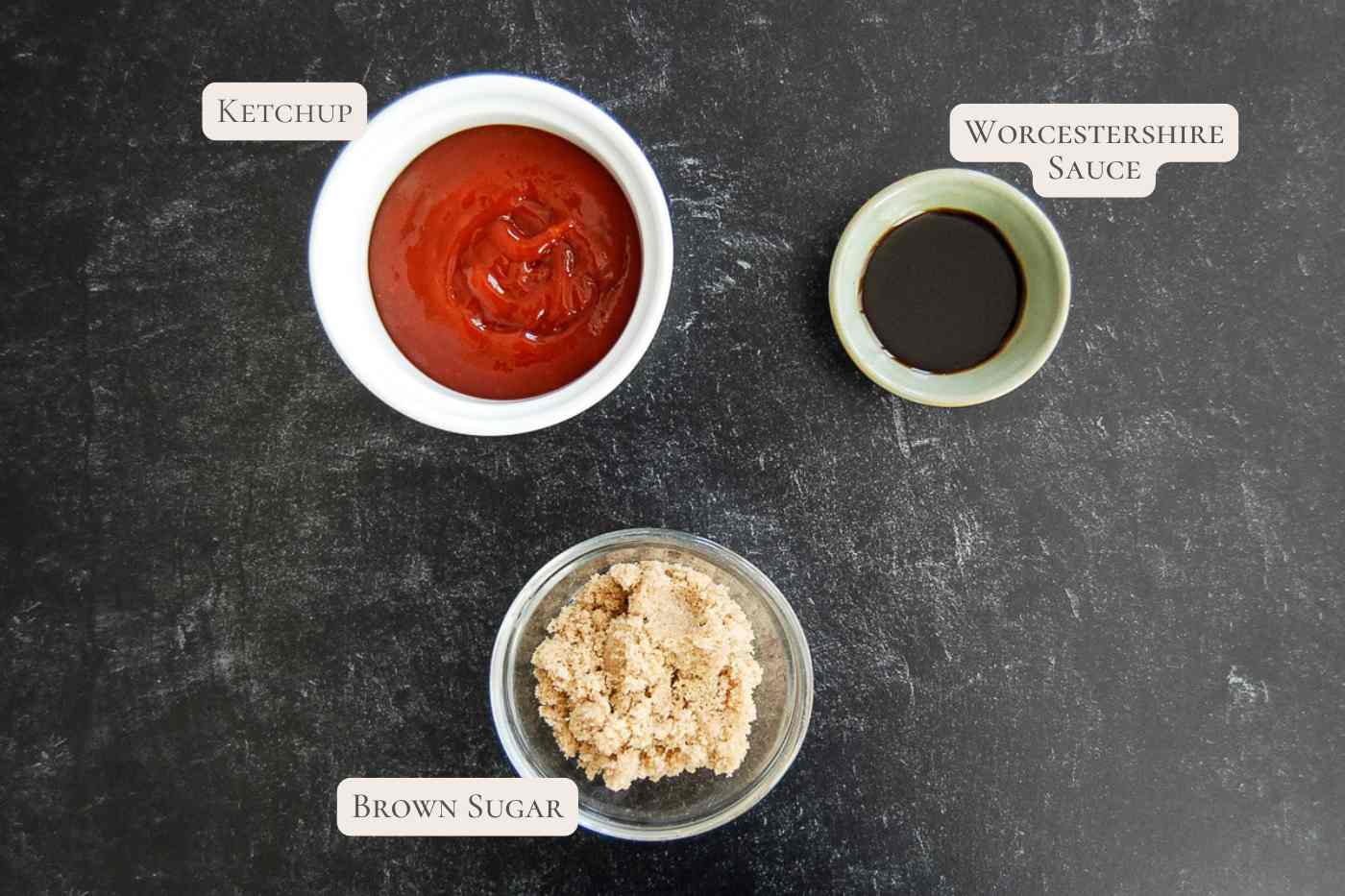 Three labeled bowls on a dark surface: one with ketchup, one with Worcestershire sauce, and one with brown sugar.