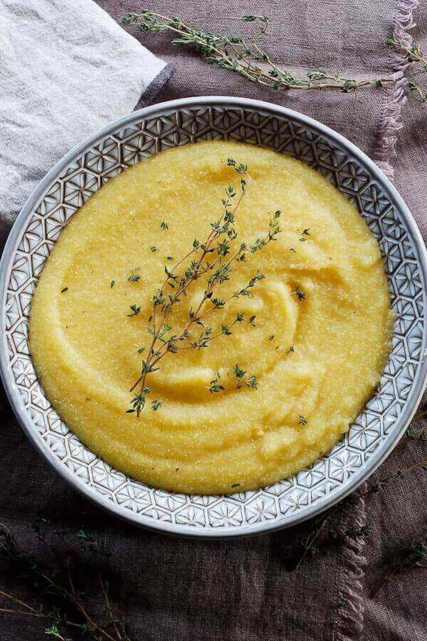 A bowl of creamy yellow polenta garnished with a sprig of fresh thyme sits on a textured brown cloth with additional thyme sprigs nearby.