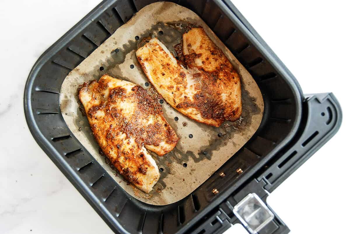 Two seasoned tilapia fillets cooked in an air fryer basket lined with parchment paper, viewed from above on a white background.