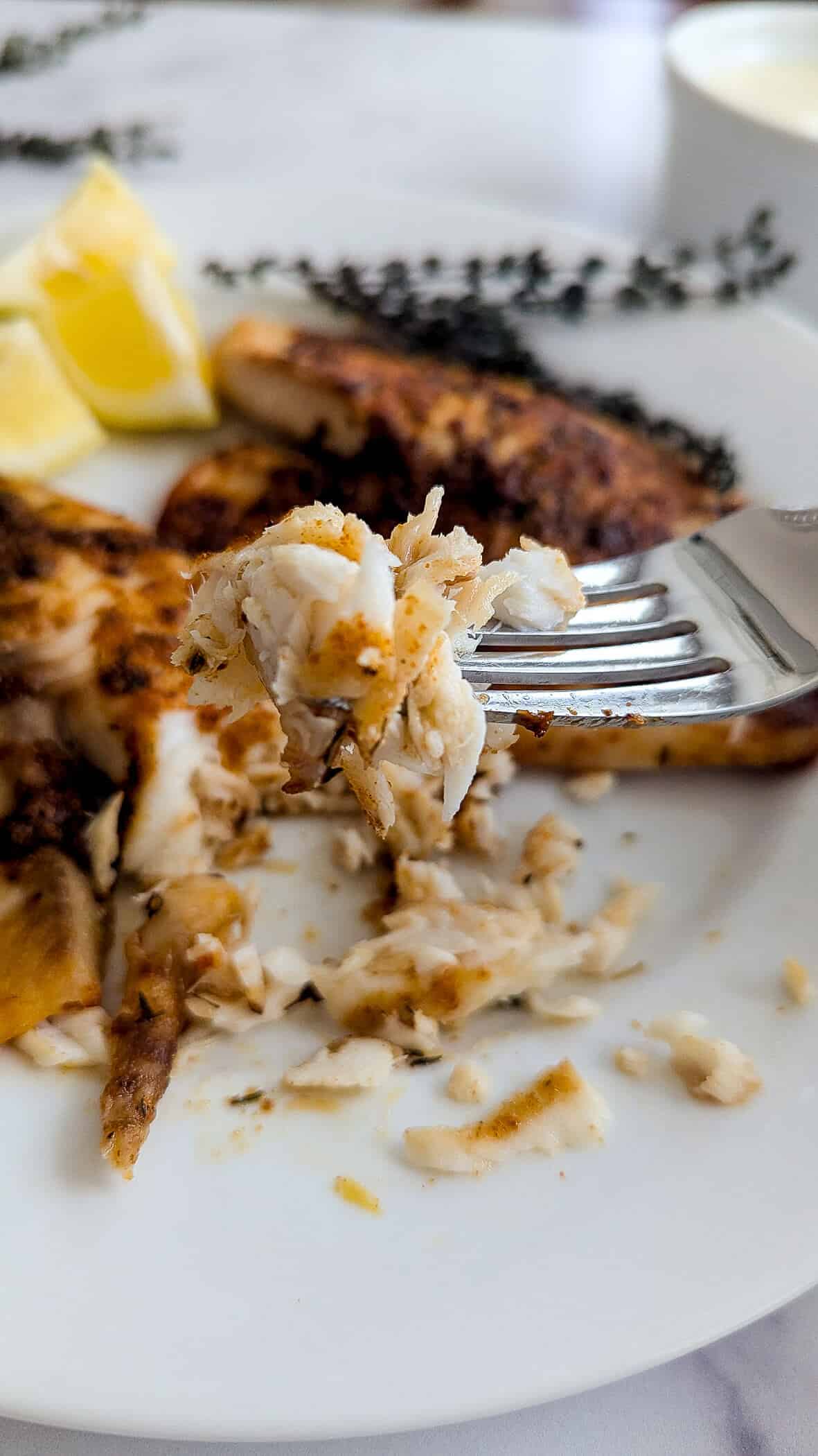 A close-up of a fork holding a bite of flaky, seasoned white fish above a plate with more tilapia fillets, lemon wedges, and a sprig of herbs in the background.