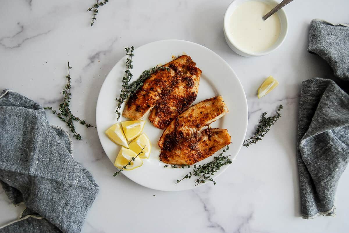 A white plate with two pieces of seasoned, air fried fish, lemon wedges, and fresh thyme sprigs on a marble surface, surrounded by gray napkins and a small bowl of white sauce with a spoon.