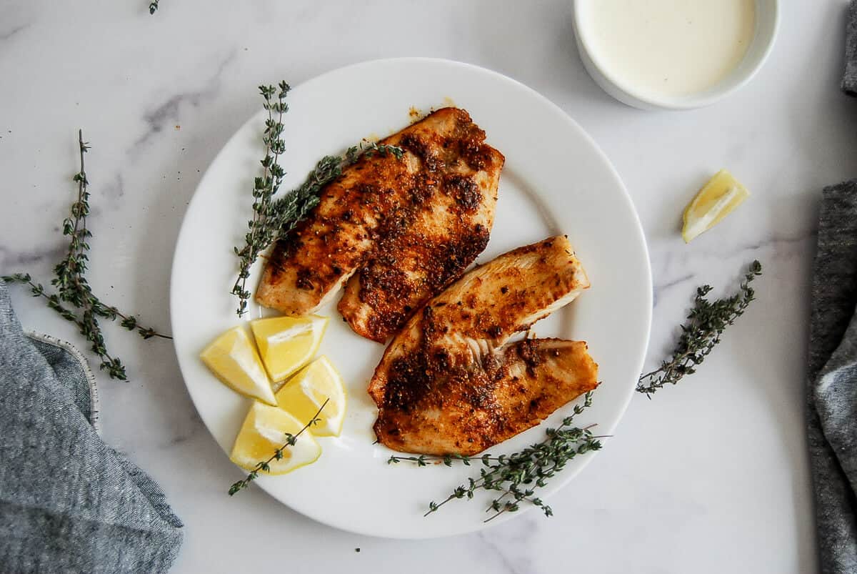 A white plate with two seasoned, cooked tilapia fillets, garnished with lemon wedges and fresh thyme sprigs, sits on a marble surface. A small bowl of white sauce and a gray cloth are nearby.
