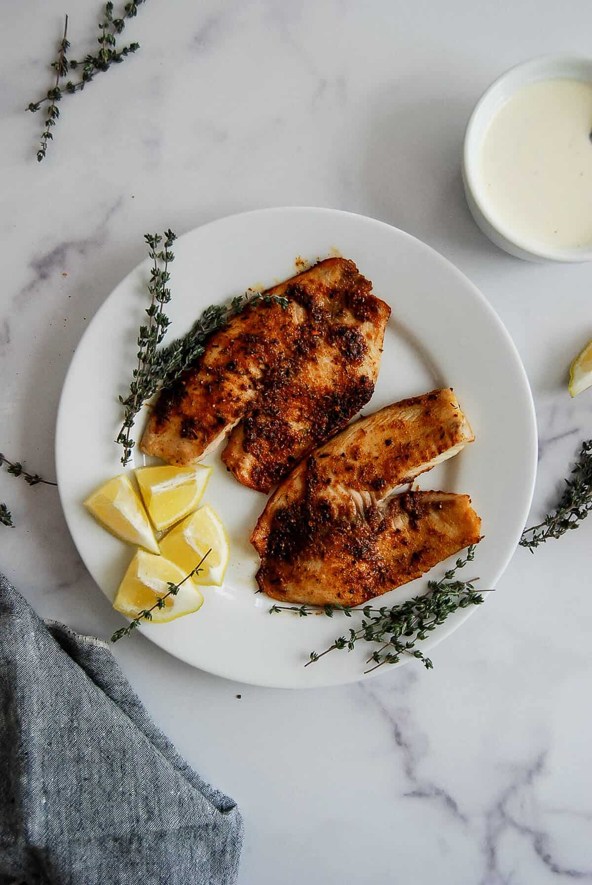A white plate with two pieces of seasoned, cooked fish, lemon wedges, and sprigs of thyme on a marble surface. A bowl of white sauce and a gray napkin are nearby.