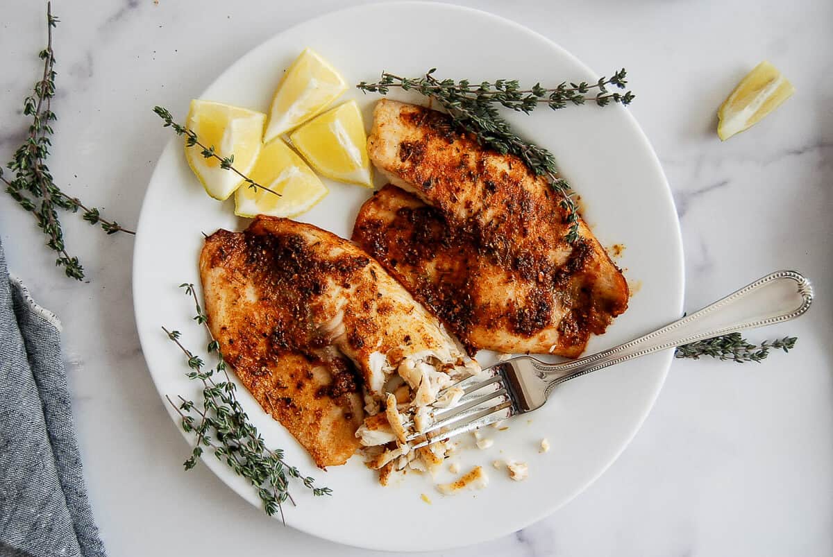 A white plate with two seasoned, air fryer tilapia fillets, garnished with fresh thyme and lemon wedges. A fork rests on the plate, flaking a portion of fish. The background is a white marble surface.