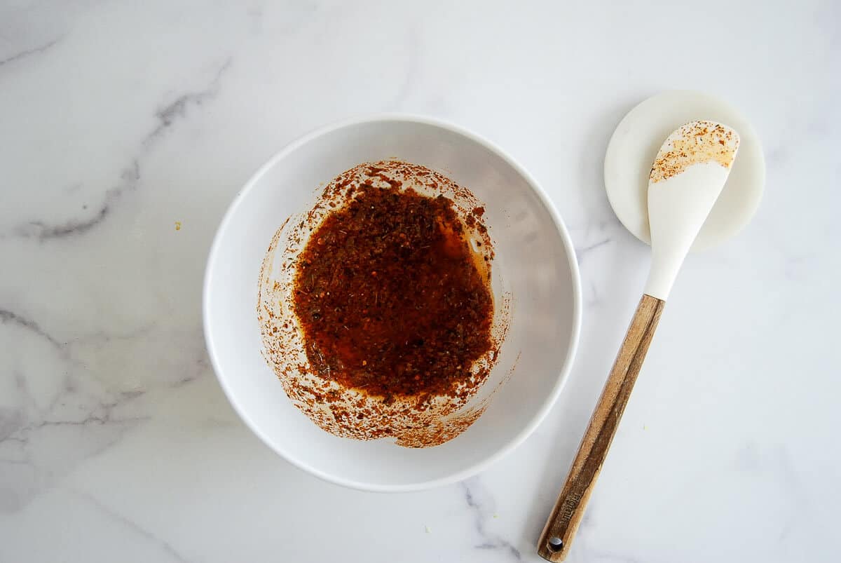 A white bowl containing a reddish-brown spice mixture sits on a white marble surface next to a white spatula with a wooden handle, which has some of the mixture on it.